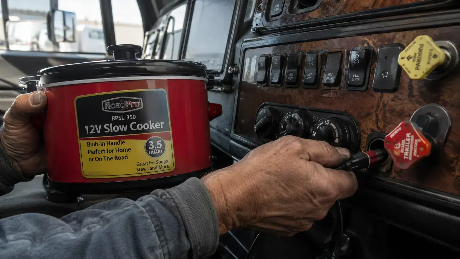 The RoadPro RPSL-350 12V slow cooker plugged into a semi truck cigarette lighter outlet, showing the cord and connector