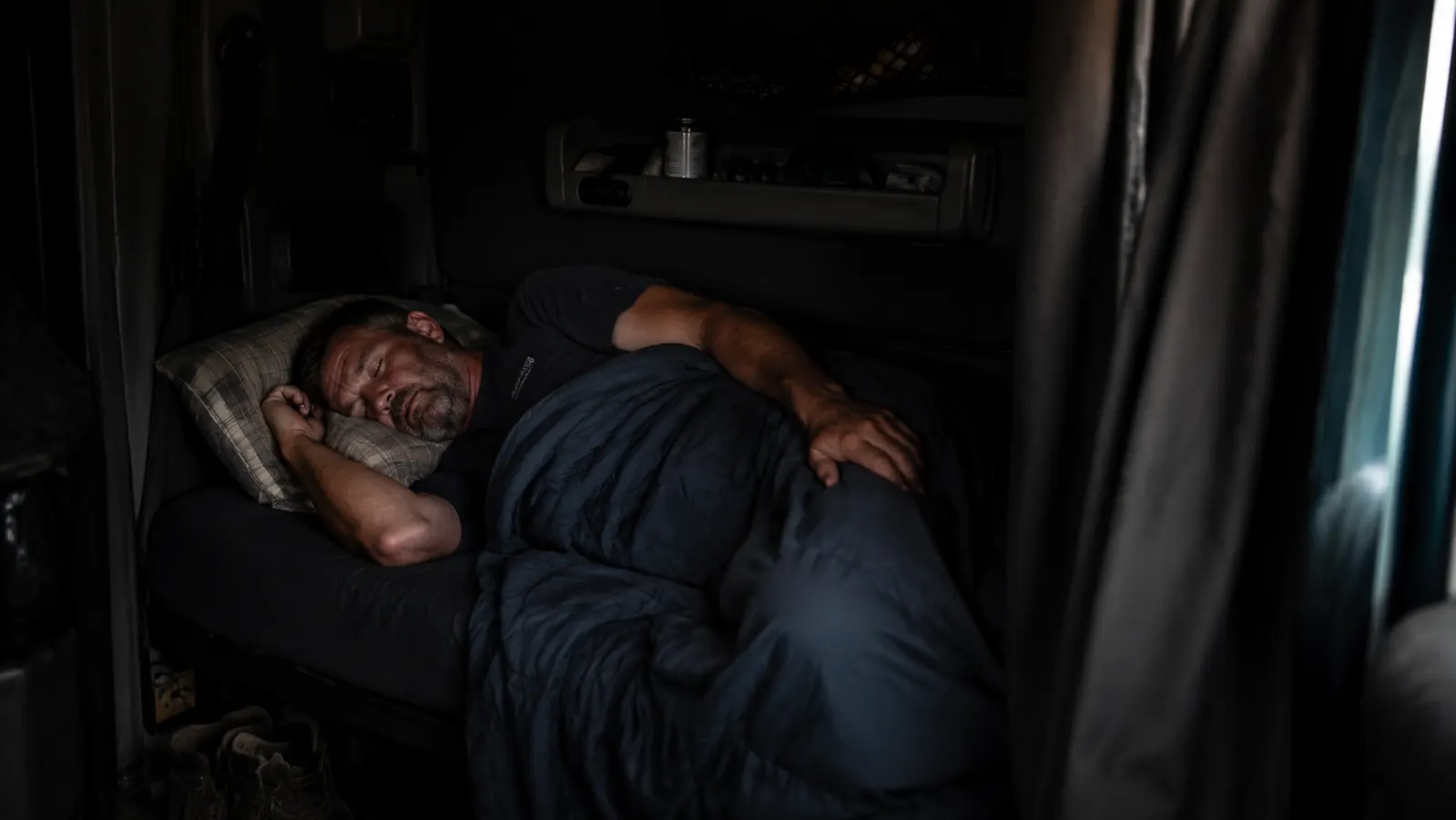 Trucker sleeping on a bunk in a well-darkened sleeper cab, arm across eyes, midday light barely visible at curtain edges