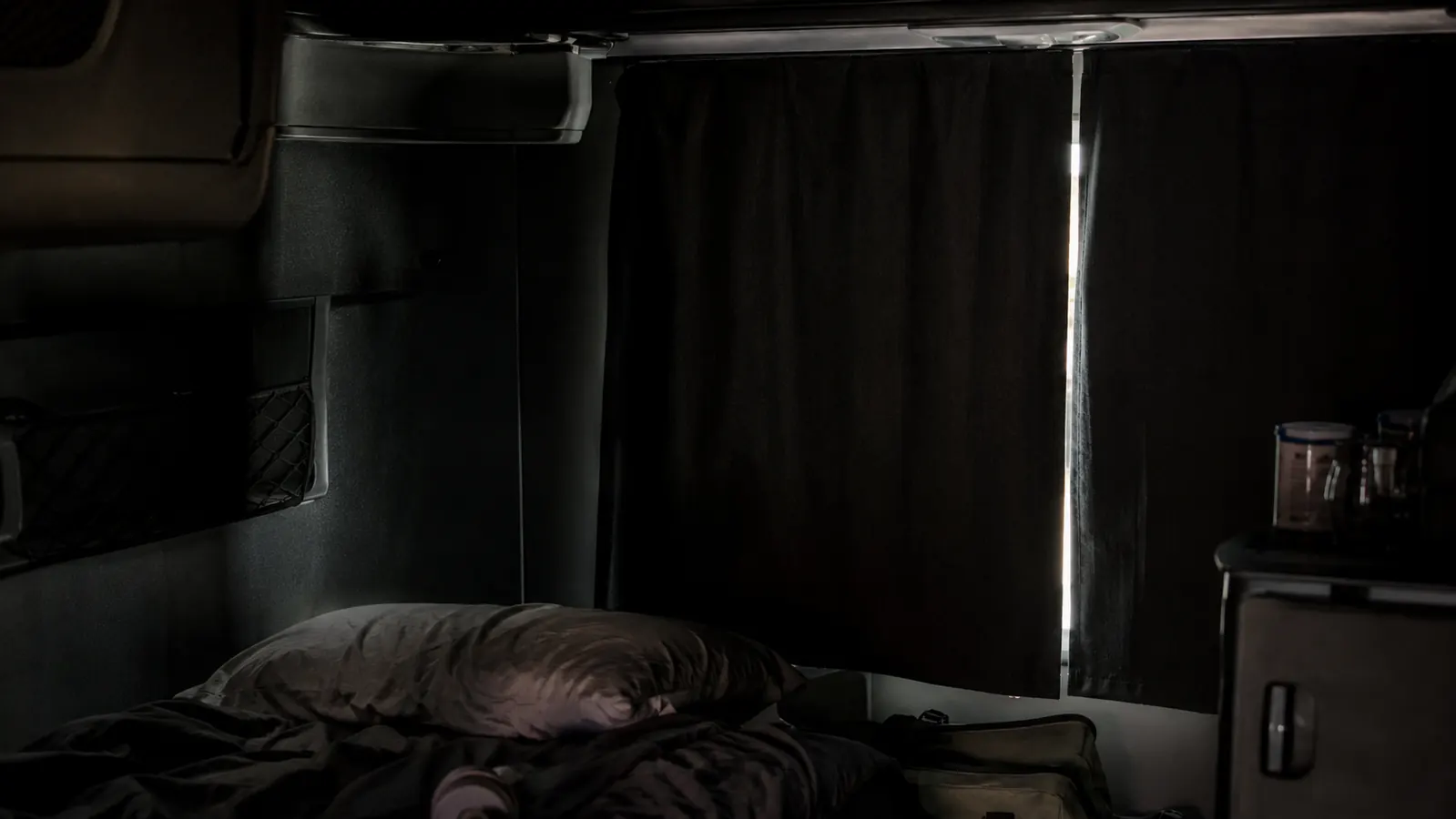 Sleeper cab interior in daytime with curtains closed, near-total darkness except for a faint line of light at the curtain edge